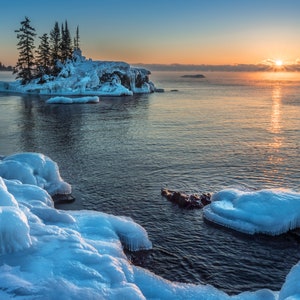 May include: A frozen lake with a small island covered in snow and trees in the foreground. The sun is setting behind the island, casting a golden glow on the water.