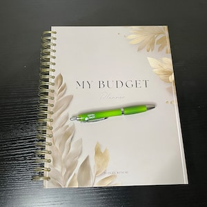 Può includere: Un quaderno beige a spirale con dettagli in foglia d'oro e il testo "MY BUDGET Planner" sulla copertina. Una penna verde è appoggiata sul quaderno. Il testo "BUDGET WITH ME" è in fondo alla copertina.