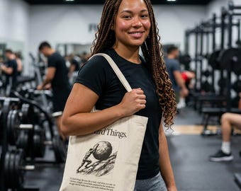May include: A beige canvas tote bag featuring a black ink illustration of a figure pushing a large boulder up a hill. The text "Hard Things." is printed above the graphic, with additional small text below the illustration on the front panel.