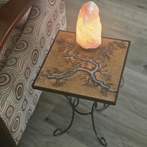 Może przedstawiać: Mały, kwadratowy stolik boczny z dekoracyjnym wzorem przypominającym drzewo na blacie. Stół ma czarną metalową ramę z zakrzywionymi nogami. Na stole stoi podświetlana, różowa lampa solna himalajska.