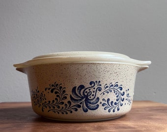 May include: A beige casserole dish with a lid, featuring a speckled pattern and blue floral designs. The dish has two handles and is suitable for baking and serving. A classic kitchenware item.