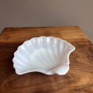 May include: A white, ceramic, seashell-shaped dish with a ribbed texture. The dish is sitting on a wooden surface.