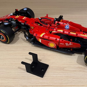 May include: A red Formula 1 race car model featuring black tires with yellow Pirelli branding, sponsor logos including HP, Shell, and Ray-Ban, and a rear wing with V-Power text. A separate black plastic stand with a U-shaped top is positioned nearby.