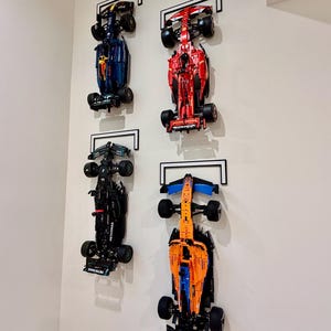 May include: Four Formula One race car models mounted vertically on a wall. The collection includes a dark blue, a bright red, a black, and an orange and blue car, each secured by a black and white rectangular frame bracket attached to the wall.