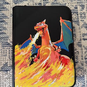 K&ouml;nnte beinhalten: Ein schwarzer, rechteckiger Ordner mit einer gemalten Illustration eines drachenartigen Wesens mit orangefarbenen Schuppen und blauen Fl&uuml;geln. Das Wesen ist mit ge&ouml;ffnetem Maul dargestellt und speit einen gro&szlig;en, leuchtenden Feuerstrahl.
