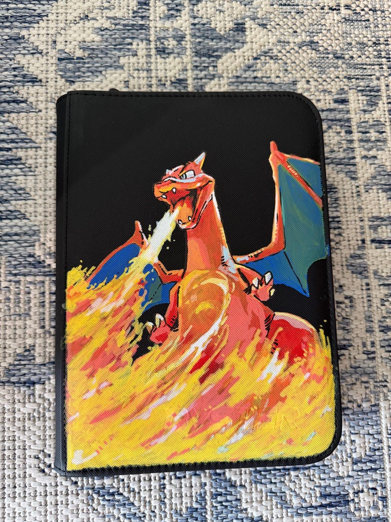K&ouml;nnte beinhalten: Ein schwarzer, rechteckiger Ordner mit einer gemalten Illustration eines drachenartigen Wesens mit orangefarbenen Schuppen und blauen Fl&uuml;geln. Das Wesen ist mit ge&ouml;ffnetem Maul dargestellt und speit einen gro&szlig;en, leuchtenden Feuerstrahl.