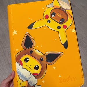 K&ouml;nnte beinhalten: Ein rechteckiger gelber Ordner mit einer aufgedruckten Illustration von zwei Charakteren, die gegenseitig ihre Kost&uuml;me tragen. Eine braune Kreatur tr&auml;gt eine gelbe Pikachu-Kapuze, eine gelbe Kreatur eine braune Evoli-Kapuze. Der Markenname Zofly ist unten rechts eingepr&auml;gt.