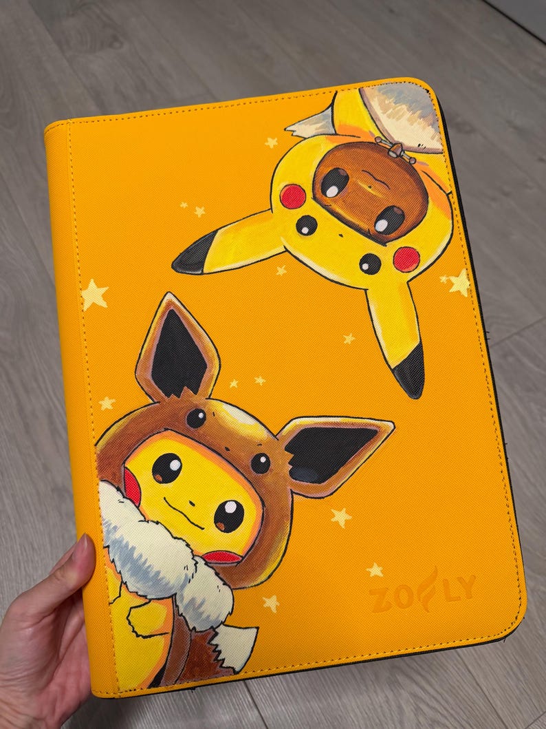 K&ouml;nnte beinhalten: Ein rechteckiger gelber Ordner mit einer aufgedruckten Illustration von zwei Charakteren, die gegenseitig ihre Kost&uuml;me tragen. Eine braune Kreatur tr&auml;gt eine gelbe Pikachu-Kapuze, eine gelbe Kreatur eine braune Evoli-Kapuze. Der Markenname Zofly ist unten rechts eingepr&auml;gt.