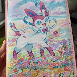 K&ouml;nnte beinhalten: Ein rechteckiger Ordner mit rosa Rand und einem aufgedruckten Bild der wei&szlig;-rosa Figur Sylveon, die in einem bunten Blumenfeld unter einem blauen Himmel steht. Das Wort SYLVEON ist unten in einer hellen Schriftart aufgedruckt.