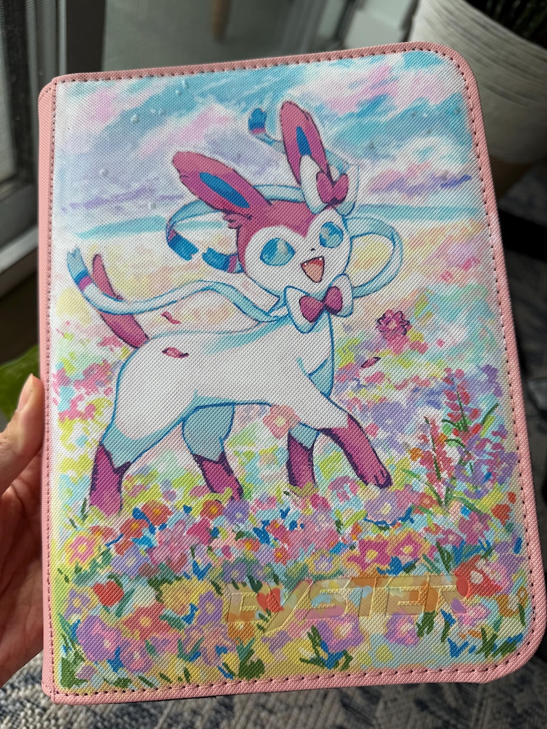 K&ouml;nnte beinhalten: Ein rechteckiger Ordner mit rosa Rand und einem aufgedruckten Bild der wei&szlig;-rosa Figur Sylveon, die in einem bunten Blumenfeld unter einem blauen Himmel steht. Das Wort SYLVEON ist unten in einer hellen Schriftart aufgedruckt.