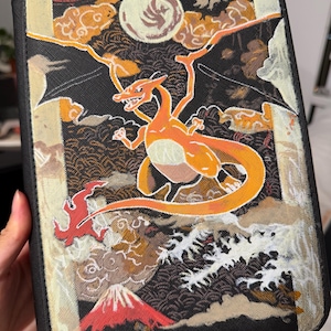 K&ouml;nnte beinhalten: Ein schwarzer, rechteckiger Ordner mit einer detaillierten Illustration eines orangefarbenen, gefl&uuml;gelten Wesens vor einem Hintergrund aus Vollmond, stilisierten Wolken, brechenden wei&szlig;en Wellen und einem roten Berggipfel.