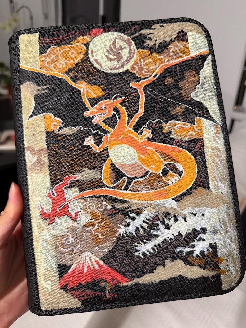 K&ouml;nnte beinhalten: Ein schwarzer, rechteckiger Ordner mit einer detaillierten Illustration eines orangefarbenen, gefl&uuml;gelten Wesens vor einem Hintergrund aus Vollmond, stilisierten Wolken, brechenden wei&szlig;en Wellen und einem roten Berggipfel.