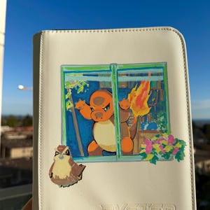 K&ouml;nnte beinhalten: Ein wei&szlig;er, rechteckiger Ordner mit einem Grafikdruck einer orangefarbenen Feuerkreatur, die in einem Fenster steht und eine Flamme am Schwanz hat, einem kleinen braunen vogelartigen Wesen darunter und dem gepr&auml;gten Text BYSTER unten rechts.
