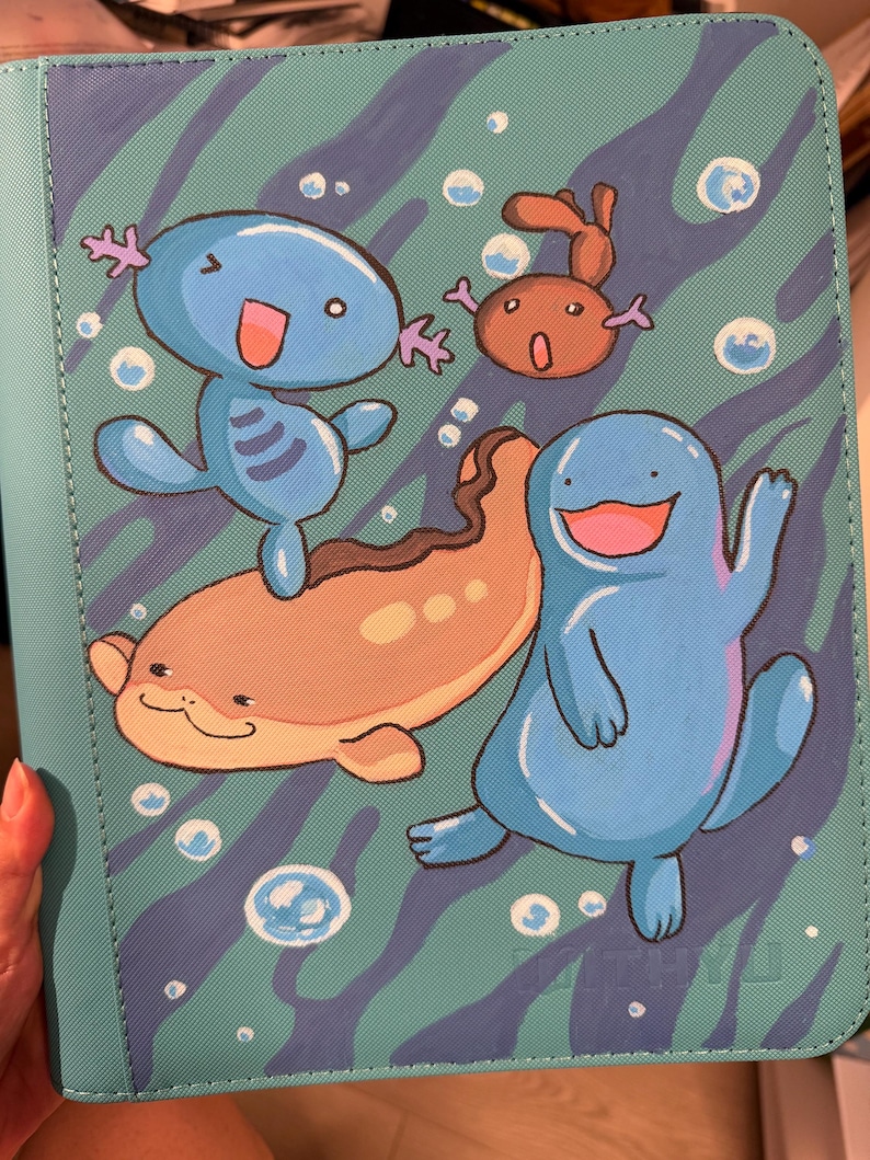 K&ouml;nnte beinhalten: Ein t&uuml;rkisfarbener Ordner mit einer gedruckten Illustration von drei blauen und braunen Wasserwesen, die zwischen wei&szlig;en Blasen schwimmen. Der Hintergrund hat ein wellenf&ouml;rmiges dunkelblaues Muster. Die Wesen umfassen eine kleine blaue Figur, eine braune und eine gr&ouml;&szlig;ere.