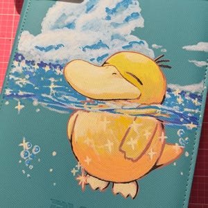 K&ouml;nnte beinhalten: Ein t&uuml;rkisfarbener, rechteckiger Ordner mit einer aufgedruckten Illustration einer gelben, enten&auml;hnlichen Kreatur, die in blauem Wasser mit Glitzereffekten schwimmt. &Uuml;ber der Kreatur ist eine gro&szlig;e wei&szlig;e Wolke vor blauem Himmel zu sehen. Unten ist BYSTER eingepr&auml;gt.