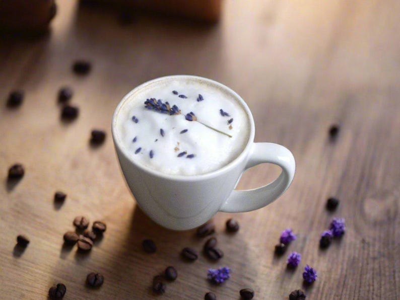 K&ouml;nnte beinhalten: Eine wei&szlig;e Keramiktasse gef&uuml;llt mit einem hellbraunen Kaffeegetr&auml;nk, das mit einer Schicht wei&szlig;en Schaums und einem Lavendelzweig gekr&ouml;nt ist.