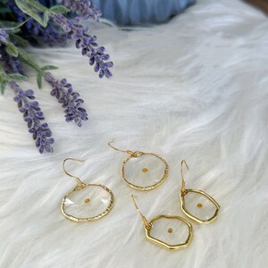May include: Gold-tone earrings with a clear resin center, each containing a mustard seed. The earrings have a unique, irregular shape and are displayed on a white, fluffy surface. Lavender sprigs are visible in the background.