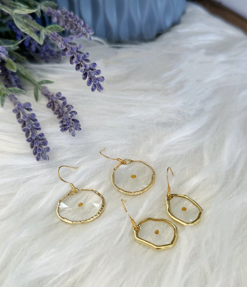 May include: Gold-tone earrings with a clear resin center, each containing a mustard seed. The earrings have a unique, irregular shape and are displayed on a white, fluffy surface. Lavender sprigs are visible in the background.