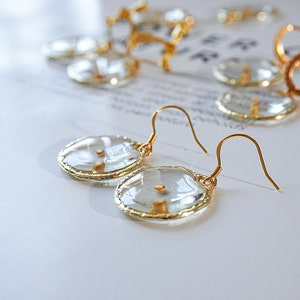 May include: Close-up of gold-colored earrings with clear, oval-shaped pendants. The pendants have a gold-colored border and a small, gold-colored detail in the center. Other gold-colored jewelry pieces are visible in the background.
