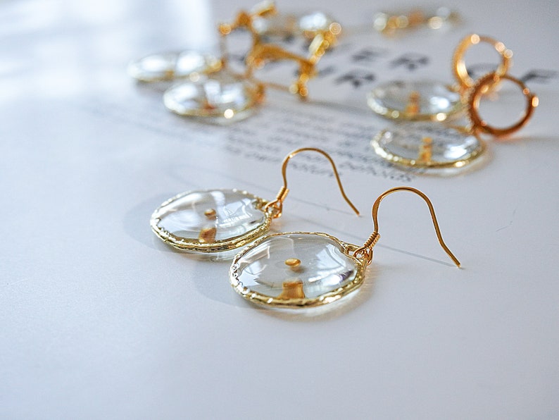 May include: Close-up of gold-colored earrings with clear, oval-shaped pendants. The pendants have a gold-colored border and a small, gold-colored detail in the center. Other gold-colored jewelry pieces are visible in the background.