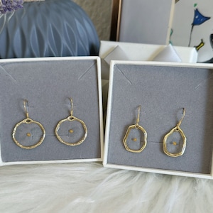 May include: Two pairs of gold-colored earrings displayed in white boxes with gray interiors. Each earring features a delicate, open-circle or irregular-shaped frame with a small, seed-like element at the center. The earrings are presented on a fluffy, white surface.