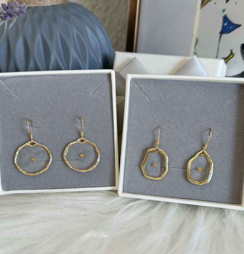 May include: Two pairs of gold-colored earrings displayed in white boxes with gray interiors. Each earring features a delicate, open-circle or irregular-shaped frame with a small, seed-like element at the center. The earrings are presented on a fluffy, white surface.