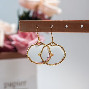 May include: A pair of gold-colored, wire-framed earrings with a clear, circular center. The earrings have a unique, irregular edge design. The earrings are hanging on a wooden display stand, with a blurred floral background.