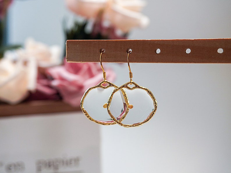 May include: A pair of gold-colored, wire-framed earrings with a clear, circular center. The earrings have a unique, irregular edge design. The earrings are hanging on a wooden display stand, with a blurred floral background.