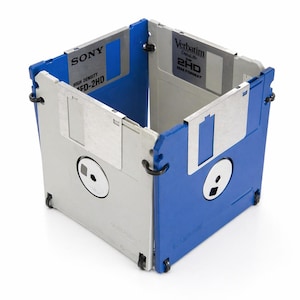 Può includere: Un contenitore a forma di cubo costruito con floppy disk da 3,5 pollici riciclati. Il contenitore presenta dischi blu e grigi, con etichette visibili di marchi come Sony e Verbatim. Il design mostra una miscela unica di tecnologia retrò e funzionalità moderna.