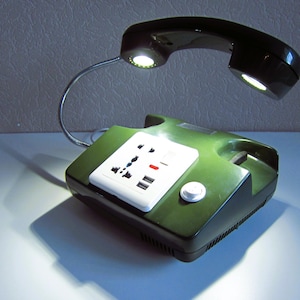 May include: A dark green desk lamp repurposed from a vintage telephone base. The handset is suspended by a flexible silver gooseneck arm, featuring two integrated LED lights. A white electrical power strip with multiple outlets is mounted on top.