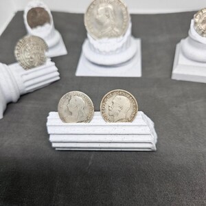 May include: Silver coins featuring profiles of historical figures, displayed on white decorative pedestals. The coins are arranged on a dark gray surface, showcasing their intricate details and historical significance. The image highlights the coins' design and the presentation.