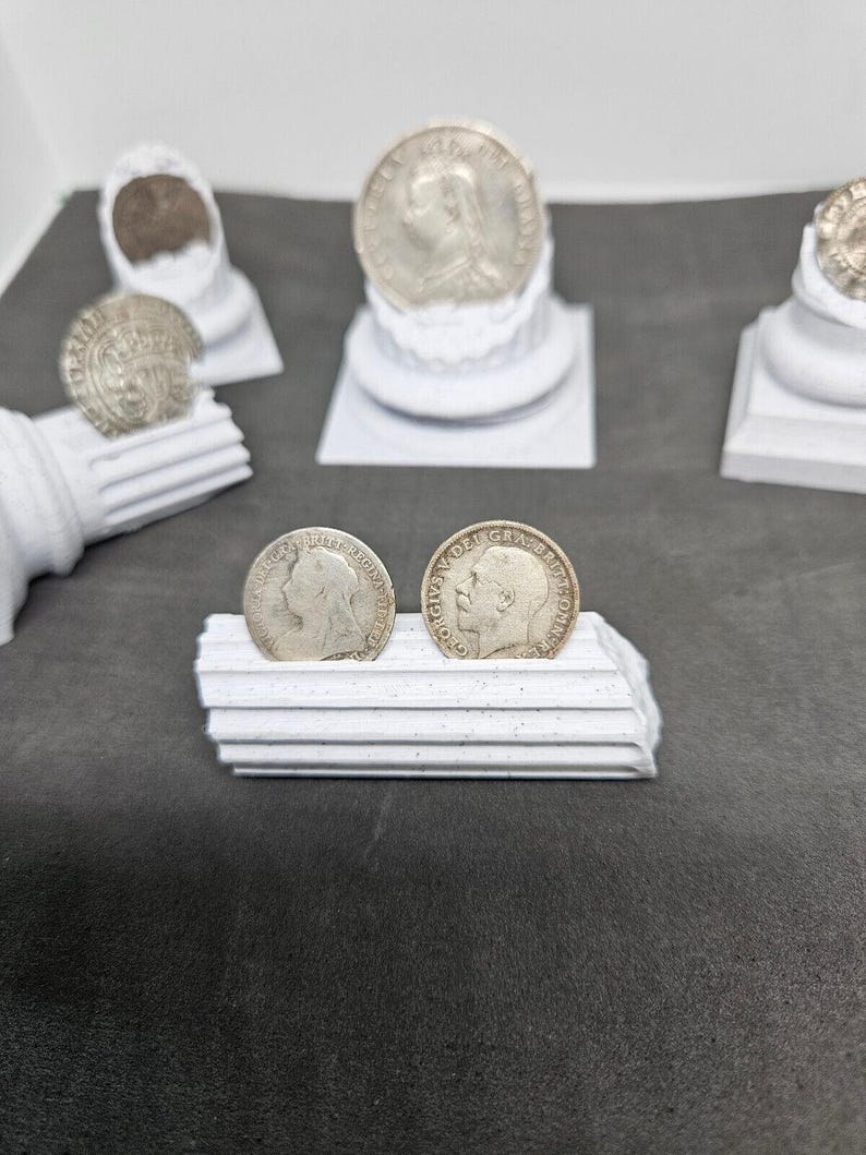 May include: Silver coins featuring profiles of historical figures, displayed on white decorative pedestals. The coins are arranged on a dark gray surface, showcasing their intricate details and historical significance. The image highlights the coins' design and the presentation.