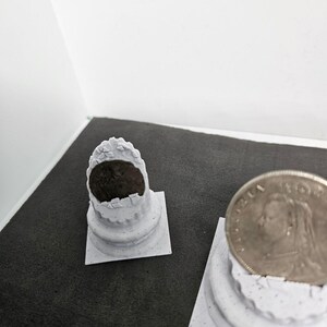 May include: Two small, white, decorative columns with a coin on top of one. The columns have a detailed, textured design. The coin features a profile of a person. The background is a dark gray surface.