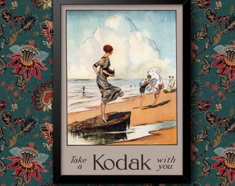 Op de afbeelding: Een vintage advertentie voor Kodak met een vrouw in een witte jurk die een camera op een strand vasthoudt met andere mensen op de achtergrond. De tekst luidt "Neem een Kodak mee."
