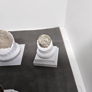 May include: A silver coin with a detailed design rests on a small, white, square-based pedestal. The coin is slightly chipped, and the pedestal has a textured surface. The background is a neutral gray and white.
