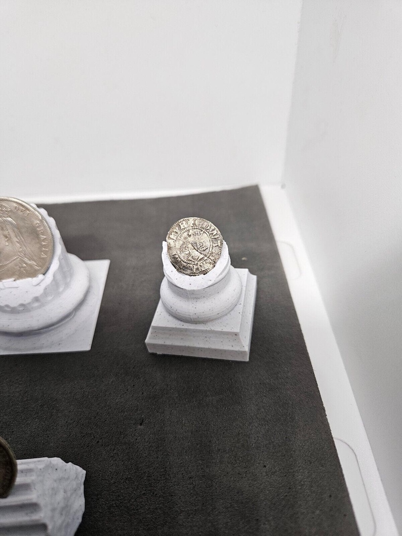 May include: A silver coin with a detailed design rests on a small, white, square-based pedestal. The coin is slightly chipped, and the pedestal has a textured surface. The background is a neutral gray and white.