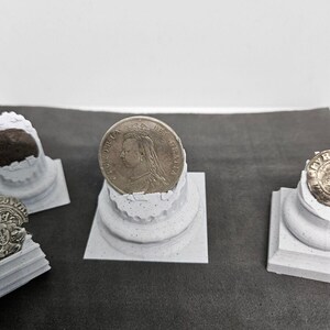 May include: A silver coin featuring a profile of a person is displayed on a small, white, decorative column. The coin is centered, with other similar columns and coins visible in the background. The columns have a textured, weathered appearance.