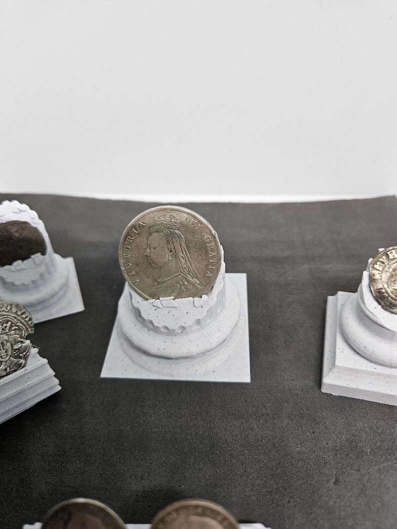 May include: A silver coin featuring a profile of a person is displayed on a small, white, decorative column. The coin is centered, with other similar columns and coins visible in the background. The columns have a textured, weathered appearance.