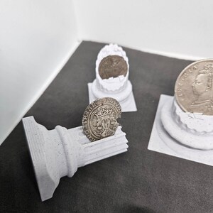 May include: A collection of antique coins displayed on small, white, 3D-printed pedestals and a column. The coins are silver-colored and feature detailed engravings. The pedestals and column are made of a speckled, light gray material.