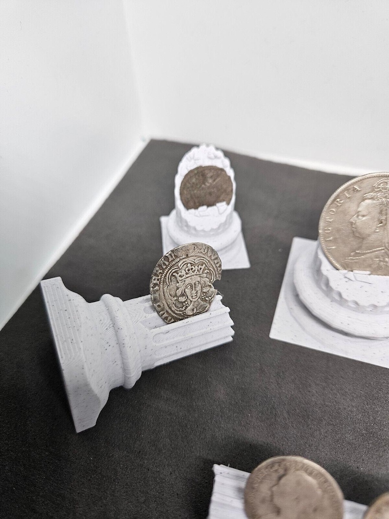 May include: A collection of antique coins displayed on small, white, 3D-printed pedestals and a column. The coins are silver-colored and feature detailed engravings. The pedestals and column are made of a speckled, light gray material.