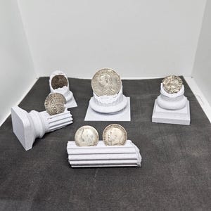 May include: A collection of antique silver coins displayed on small, white, architectural-style pedestals. The coins feature detailed portraits and inscriptions. The arrangement includes coins of various sizes and designs, showcasing historical numismatic pieces.