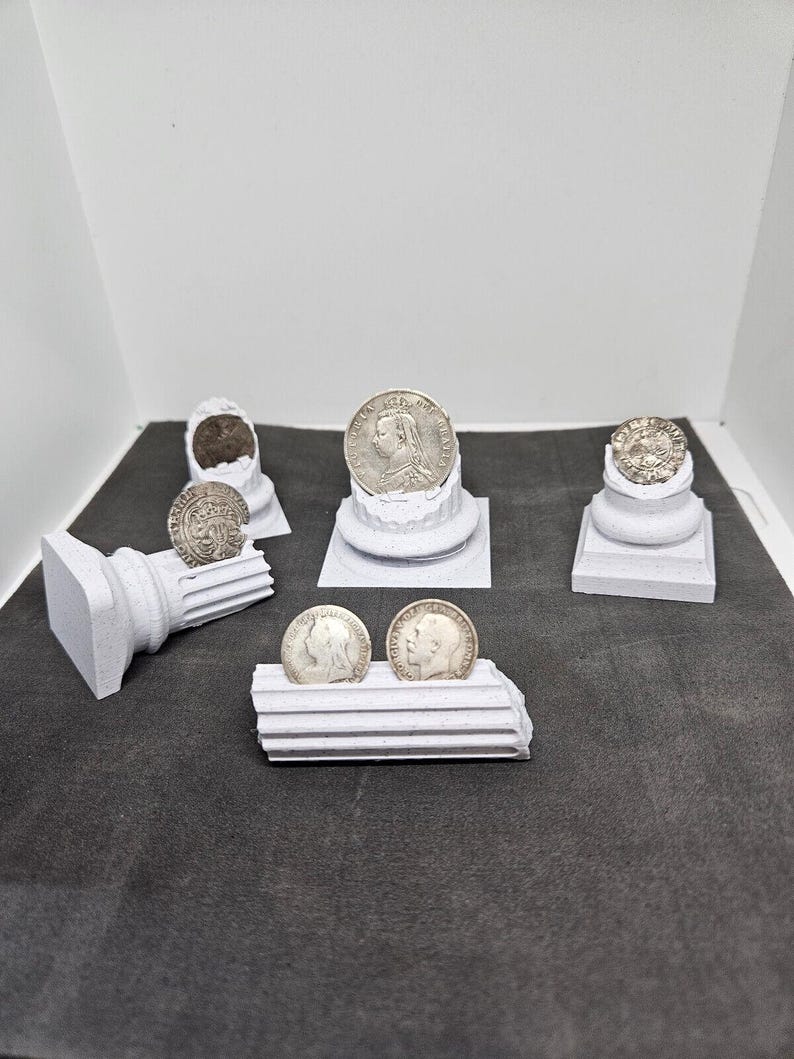 May include: A collection of antique silver coins displayed on small, white, architectural-style pedestals. The coins feature detailed portraits and inscriptions. The arrangement includes coins of various sizes and designs, showcasing historical numismatic pieces.