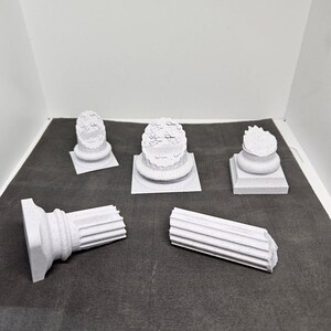 May include: A collection of small, white, architectural column models. The models include complete columns and broken column sections, showcasing various classical designs. The pieces are arranged on a dark gray surface.