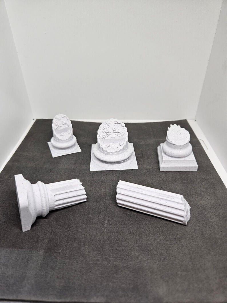 May include: A collection of small, white, architectural column models. The models include complete columns and broken column sections, showcasing various classical designs. The pieces are arranged on a dark gray surface.