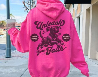 Op de afbeelding: Een felroze hoodie met een trekkoord. De achterkant van de hoodie toont de tekst "Unleash Your Faith" met een afbeelding van een hond en de zinnen "Pray More" en "Worry Less".