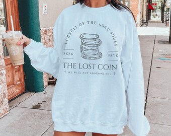 Op de afbeelding: Een lichtblauw sweatshirt met een grafische print van een stapel munten en de tekst "Pursuit of the Lost Souls", "Seek", "Save", "The Lost Coin" en "He will not abandon you".
