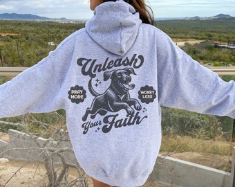 Op de afbeelding: Grijs hoodie met een zwarte hond grafische en de tekst "Unleash Your Faith" met de woorden "Pray More" en "Worry Less" aan weerszijden van de hond.