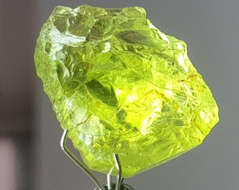 May include: A vibrant, translucent, lime-green gemstone with a rough, textured surface. The gem is held by a silver metal stand, showcasing its natural, irregular shape. The stone's color and clarity suggest it is a peridot, a popular choice for jewelry.