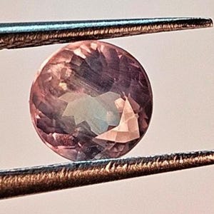 May include: A round, faceted gemstone held by tweezers. The gem has a mix of pink, purple, and blue hues. The tweezers are silver-colored and the background is a soft, neutral tone.