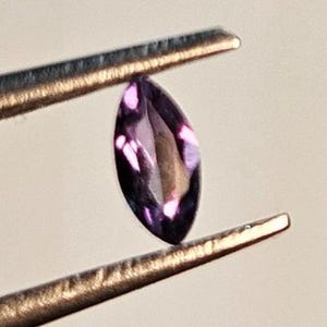 May include: A marquise-cut, deep purple gemstone is held by a pair of tweezers. The gemstone has a polished surface and a pointed, oval shape. The tweezers are made of metal and have a textured surface.